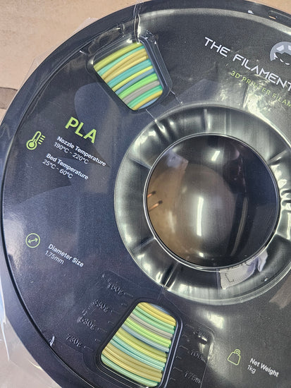 Spool of filament labeled 'The Filament' with PLA material on a neutral background